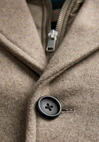 Brun uldblazer med en hakket krave, en sort knap og en sølvlynlås. Teksturen virker blød og fleece-agtig.