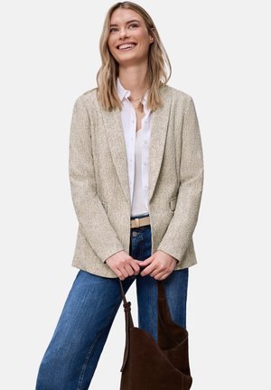 Lachende vrouw die een beige gestructureerd colbert draagt, een wit overhemd met knopen, blauwe spijkerbroek en met beide handen een donkerbruine leren handtas vasthoudt.