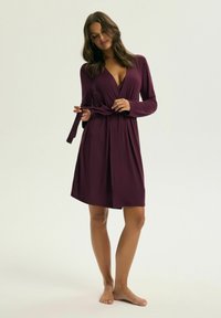 Bordowa sukienka kopertowa z długimi rękawami, dekoltem w serek i paskiem w talii. Gładki materiał i długość do kolan, o luźnym kroju.