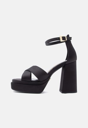Crna platforma sandala s visokim potpeticama, širokim ukriženim remenčićima i remenom oko gležnja s zlatnom kopčom.