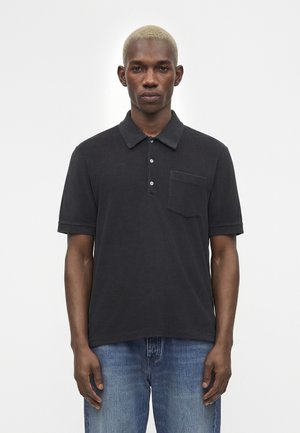 Jeune homme aux cheveux blonds courts portant un polo noir à manches courtes avec une poche poitrine et un jean bleu, debout devant un fond uni.