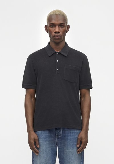 Jeune homme aux cheveux blonds courts portant un polo noir à manches courtes avec une poche poitrine et un jean bleu, debout devant un fond uni.