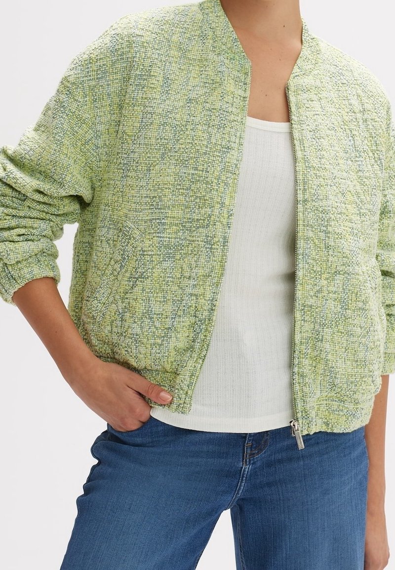 Personne portant une veste zippée vert clair texturée sur un débardeur blanc côtelé et un jean bleu, avec une main dans une poche.