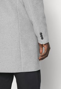 Ciemnoskóra dłoń spoczywająca obok rękawa jasnoszarego wełnianego płaszcza z trzema czarnymi guzikami, zestawiona z ciemnymi spodniami na jednolitym tle.