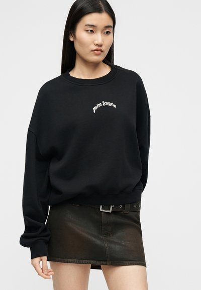 Νεαρή γυναίκα με μακριά ίσια μαλλιά, που φοράει μαύρο φούτερ «Palm Angels» και κοντή σκούρα δερμάτινη φούστα με ζώνη.