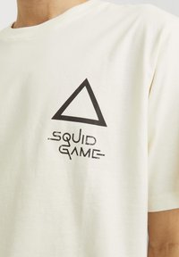 Camiseta de algodón color crema con un gráfico triangular negro y el texto "SQUID GAME" debajo en una fuente audaz y desgastada. Mangas cortas, cuello redondo.