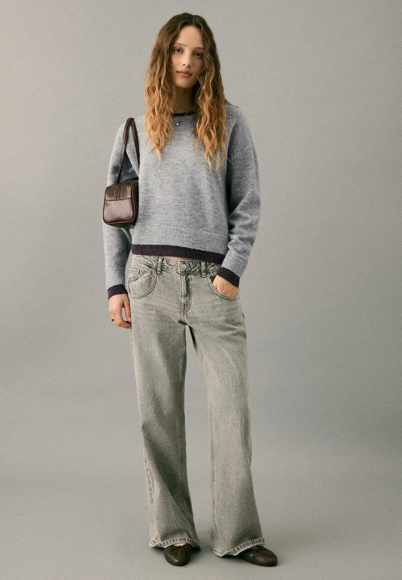 Sivi pleteni džemper s tamnim rebrastim rubom, uparen sa svijetlosivim trapericama širokih nogavica. Smeđa torba preko ramena i tamna obuća upotpunjuju odjevnu kombinaciju.