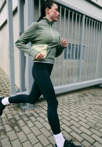 O fato atlético apresenta um casaco de fecho com textura em verde-oliva, leggings pretas e um saco em amarelo claro. Correndo numa calçada de tijolos.