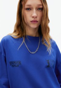 Hanorac albastru supradimensionat cu logo-ul "Stranger Things" și un alt logo text negru pe piept. Colier din lanț argintiu completează ținuta.