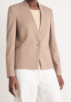 Kvinde iført en beige skræddersyet blazer med en enkelt knap og hvide bukser, stående mod en ensartet lys baggrund.