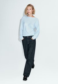 Donna che cammina con le mani nelle tasche, indossando un maglione azzurro chiaro, jeans a gamba larga scuri e scarpe nere, su uno sfondo bianco semplice.