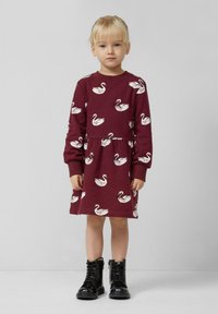 Maronfarbendes Kleid mit weißen Schwänen, langen Ärmeln, rundem Halsausschnitt und geraffter Taille. Kombiniert mit schwarzen Lackstiefeletten bis zum Knöchel.