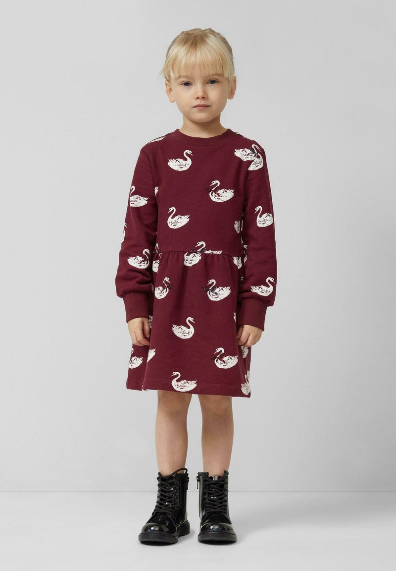 Maronfarbendes Kleid mit weißen Schwänen, langen Ärmeln, rundem Halsausschnitt und geraffter Taille. Kombiniert mit schwarzen Lackstiefeletten bis zum Knöchel.