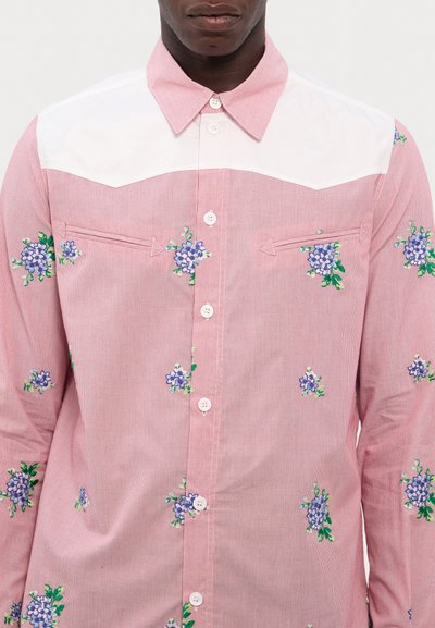 Chemise rayée rouge et blanche avec broderie florale. Présente un empiècement blanc, un devant boutonné et deux poches poitrine avec rabats pointus.