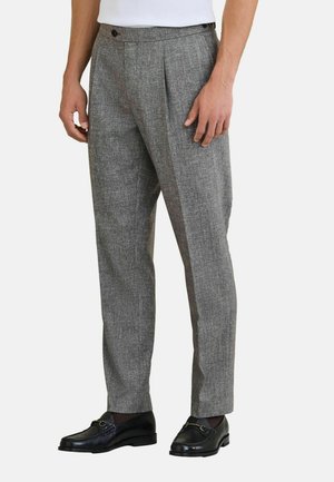 Mann trägt graue, maßgeschneiderte Hose mit Falten und schwarze Lederloafers, steht vor einem schlichten hellen Hintergrund.