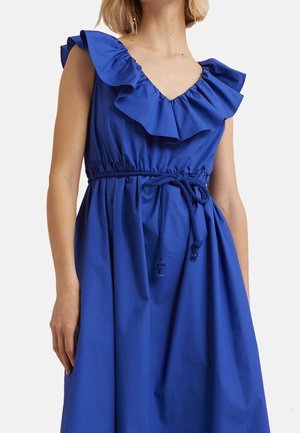 Kobieta ubrana w niebieską sukienkę bez rękawów z falbanianym dekoltem w kształcie litery V oraz plecionym paskiem zawiązanym w talii.
