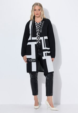 Langer schwarzer Cardigan mit weißen geometrischen Mustern, versehen mit dem Text "MODERN" und "VIEW." Kombiniert mit schwarzen Lederhosen und weißen Absätzen.