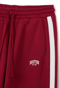 Pantalones de chándal burdeos con una franja lateral blanca. Cuenta con una cinturilla elástica y el logo bordado "BOSTON USA 1891" en blanco. Textura de tela suave.