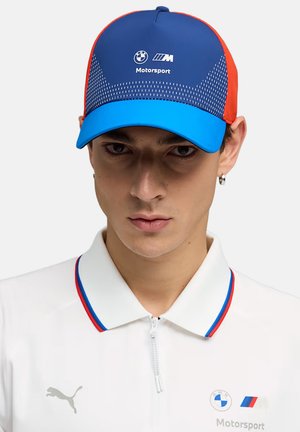 Giovane uomo che indossa un cappellino BMW Motorsport blu e rosso e una polo bianca con zip con il logo Puma e gli emblemi BMW Motorsport.