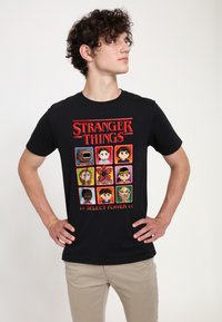 Camiseta negra de algodón con arte pixelado de "Stranger Things", que muestra nueve retratos de personajes y el texto "SELECT PLAYER" en rojo.