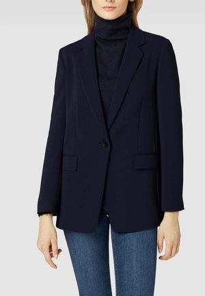 Žena ve tmavě modrém blejzru přes černý rolák a modré džíny, stojící před jednobarevným světlým šedým pozadím.