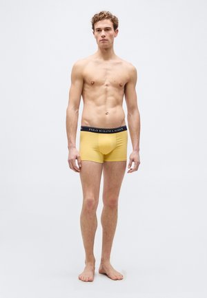 Jeune homme debout pieds nus sur fond blanc portant un boxer Polo Ralph Lauren jaune.