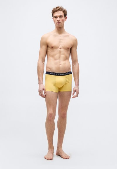 Jeune homme debout pieds nus sur fond blanc portant un boxer Polo Ralph Lauren jaune.