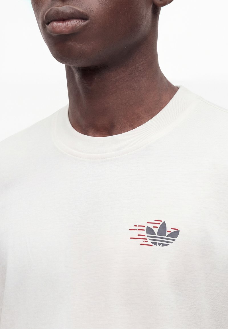 Primo piano di una persona che indossa una camicia bianca con un piccolo logo a foglia stilizzato nero e rosso sul petto.