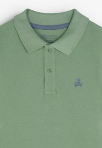 Polo vert en tissu texturé. Il présente un col classique, deux boutons et un petit emblème de crâne bleu sur la poitrine.