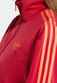 Röd zip-jacka med hög krage, som har tre orange ränder på axeln och en gul logotyp på bröstet. Mjukt, atletiskt tyg.