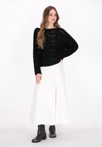 Schwarzer gestrickter Pullover mit lockerer Passform und offener Musterung, kombiniert mit einem weißen Maxirock und schwarzen Ankle Boots mit einem Riemenakzent.