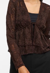 Femme portant un chemisier en dentelle florale marron foncé à manches longues avec un nœud à l'avant, sur un haut noir et un pantalon noir.