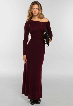 Mujer con un vestido largo de hombros descubiertos color rojo oscuro, sosteniendo un bolso negro con flecos, parada frente a un fondo liso y claro.