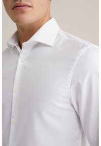 Baltas marškinių tipo palaidinė su struktūruota apykakle, viena sagtele, lygiu audiniu ir subtiliu blizgesiu. Figūrai pritaikyta forma, be raštų ar akcentų.