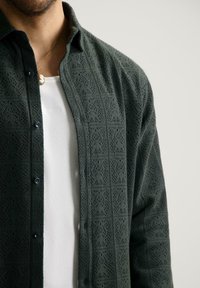 Man in een donkergroene gestructureerde open shirt over een witte geribbelde tanktop, met een gouden camee-hangertje.