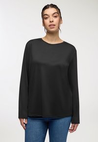 Camicetta nera a maniche lunghe in un tessuto morbido e leggero con scollatura tonda. Abbinata a jeans in denim blu. Texture liscia senza motivi.