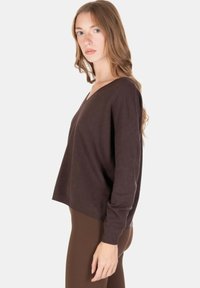 Donna con capelli lunghi castano chiaro che indossa un maglione scollo a V ampio di colore marrone scuro e pantaloni marroni aderenti, in piedi contro uno sfondo chiaro.