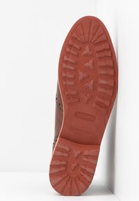 Semelle de chaussure habillée en cuir marron avec un motif de bande de roulement et le nom de la marque embossé sur la semelle extérieure en caoutchouc, inclinée contre un fond blanc d'angle.