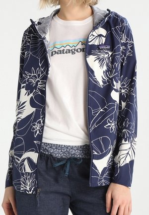 Person iført en hvid t-shirt med Patagonia-logo, navyblå blomstret lynlåstrøje og mørkeblå bukser med snøre.