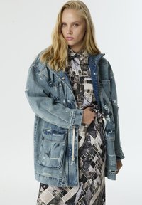 Blonde vrouw in een bedrukte jurk en een oversized versleten spijkerjack, die met één hand het jack bij de taille vasthoudt tegen een witte achtergrond.