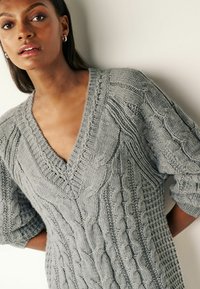 Maglione grigio a maglia con scollatura a V, intricati motivi a treccia e maniche corte e ampie. Il tessuto appare texture e morbido.