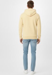 Sudadera con capucha de color amarillo claro, con capucha grande, textura suave y puños acanalados, combinada con vaqueros azul claro y zapatillas blancas. Vista trasera.