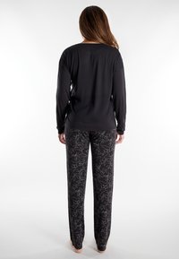 Woman with long hair stands barefoot, wearing a black long-sleeve top and black pants with a white floral line pattern, shown from the back.