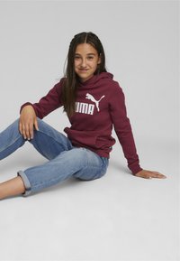 Sudadera color marrón con un logo blanco de "PUMA" y la silueta de un gato, combinada con unos jeans claros de tiro bajo y enrollados. Textura suave y corte casual.