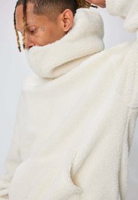 Pull-over en polaire blanche avec col montant, tissu texturé et poches latérales. Présente une coupe ample et un design décontracté.