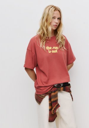 Oversized rode t-shirt met gele tekst "de zon schijnt," gedragen met witte broek en een multicolor gebreide omslag. Zwarte riem met ronde gesp.