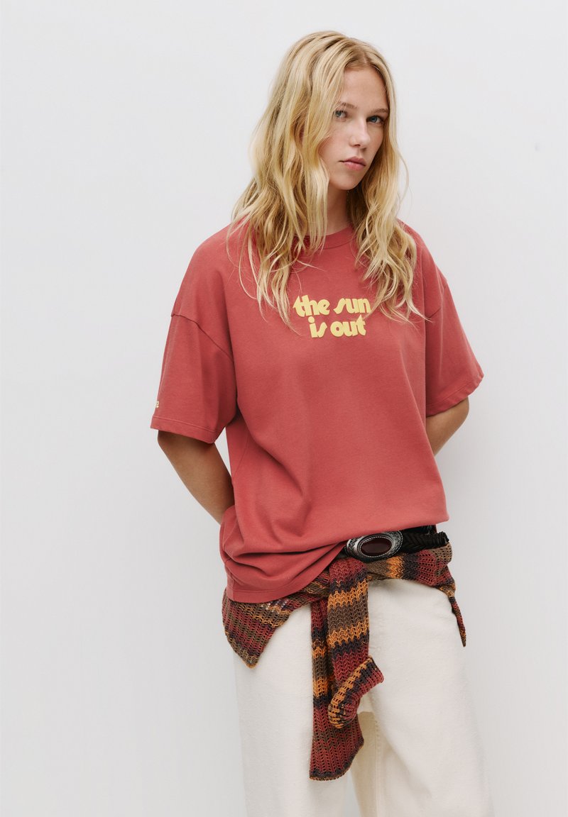 Übergroßes rotes T-Shirt mit gelbem Text "die Sonne scheint", getragen mit weißen Hosen und einem mehrfarbigen gestrickten Schal. Schwarzer Gürtel mit runder Schnalle.