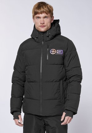 Man wearing black padded winter jacket with hood and logo on chest, standing against a plain light background.