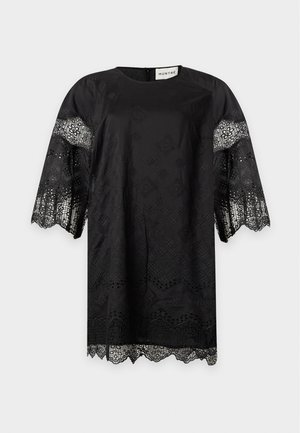 Femme portant une robe en dentelle noire avec des manches larges et des bords festonnés, associée à des escarpins pointus de couleur claire, se tenant sur un fond gris.