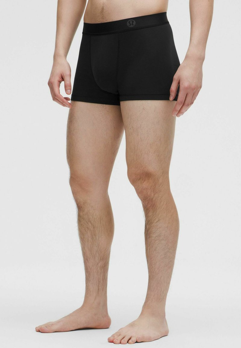 Melnas bokseršortus ar gludu, elastīgu audumu, ar platu jostasvītru, kurai priekšā ir logs, izgriezti, lai veidotu pieguļošu formu.